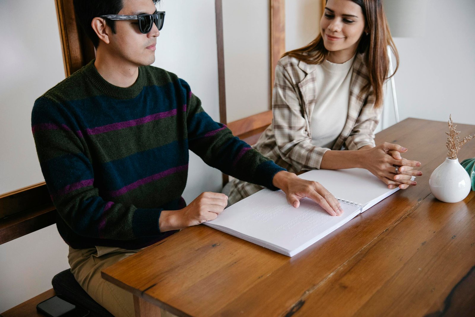 A visually impaired man reads a Braille book with a supportive woman sitting beside him indoors.
